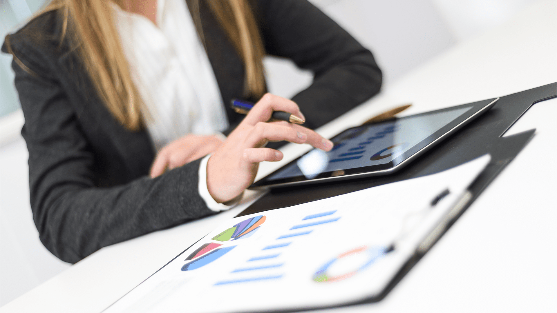 A person dressed in a business suit is interacting with a tablet, tapping on a graph or chart displayed on the screen. In front of them on the desk are printed documents with colorful pie charts and bar graphs. A pen is held in their other hand, and the setting appears to be a professional office environment.