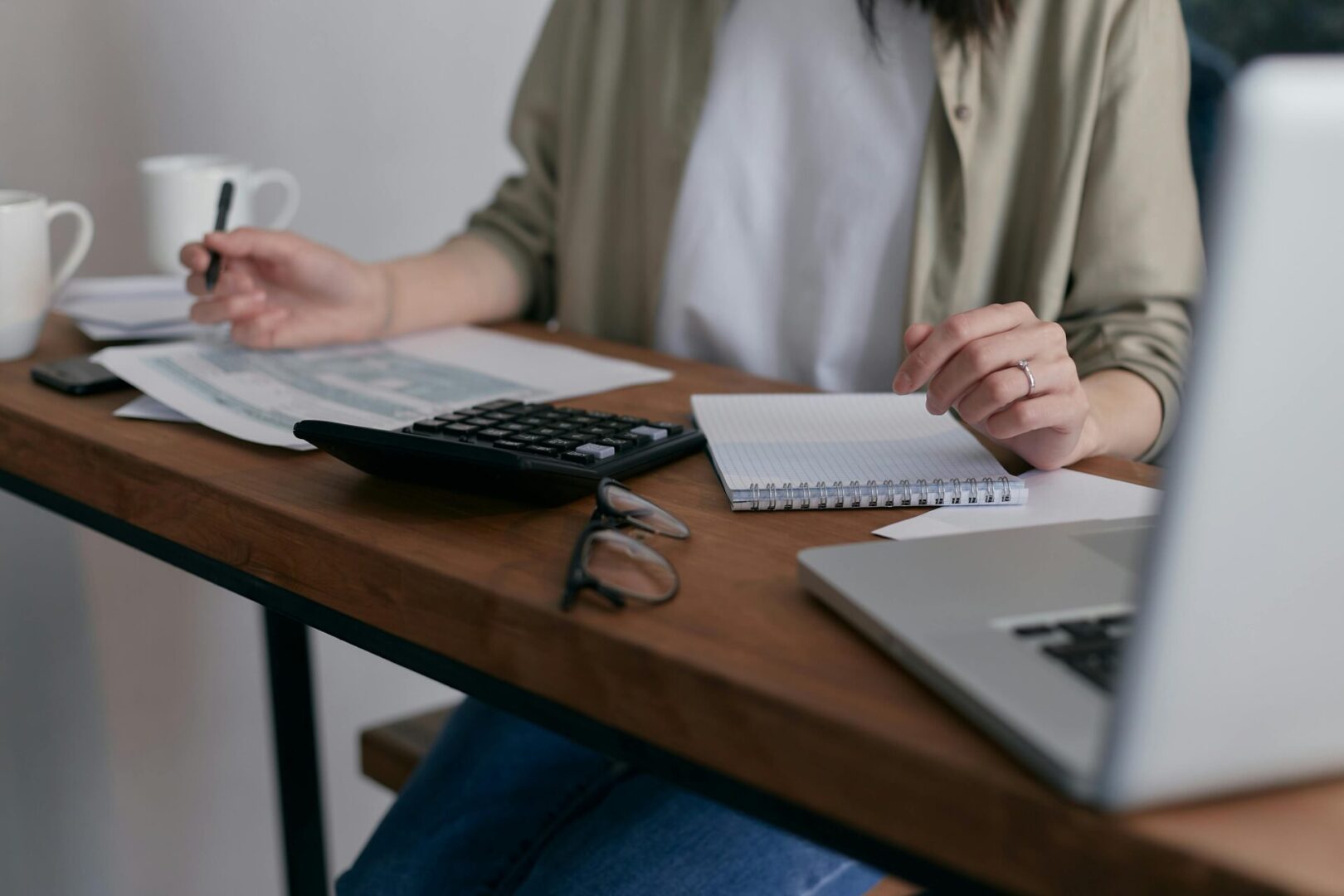 Person sitting at a desk with papers, a calculator, a notebook, glasses, and a laptop, appearing to work or study.
