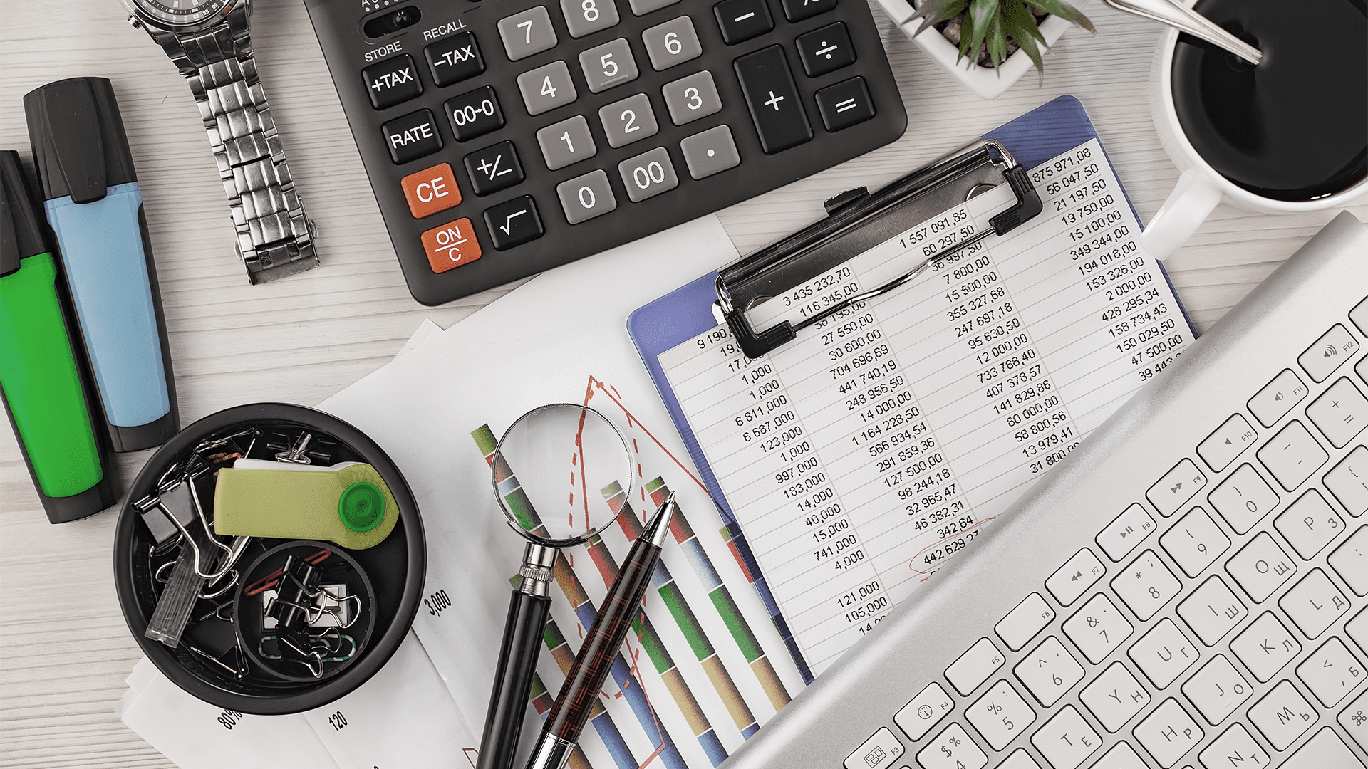 A cluttered office desk with various tools and paperwork. A calculator, a wristwatch, and highlighters in green, blue, and black are scattered across the desk. There are also paperclips, a USB drive, a magnifying glass, and two pens. In the background, financial documents with bar graphs and tables are visible. A clipboard holds a spreadsheet, and a laptop keyboard is partially shown. A small plant and a cup of coffee are visible in the top-right corner of the image.