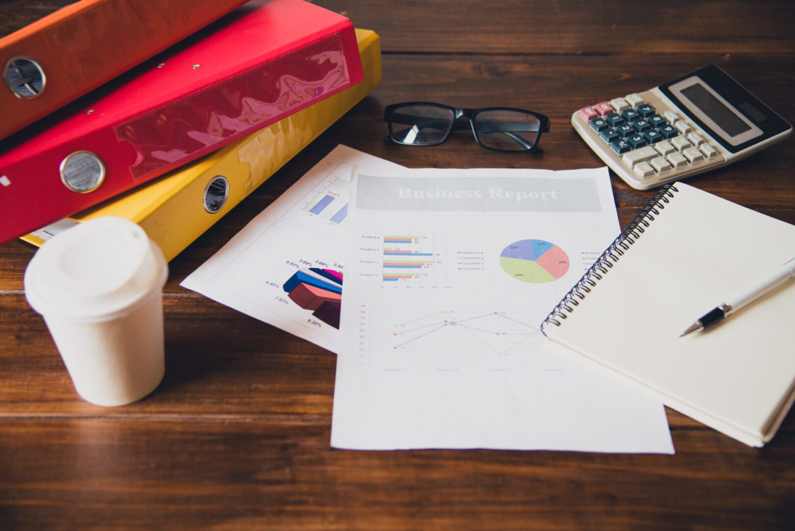 Desk with business reports, colorful binders, a calculator, notebook, glasses, and a coffee cup, representing office work or accounting tasks.