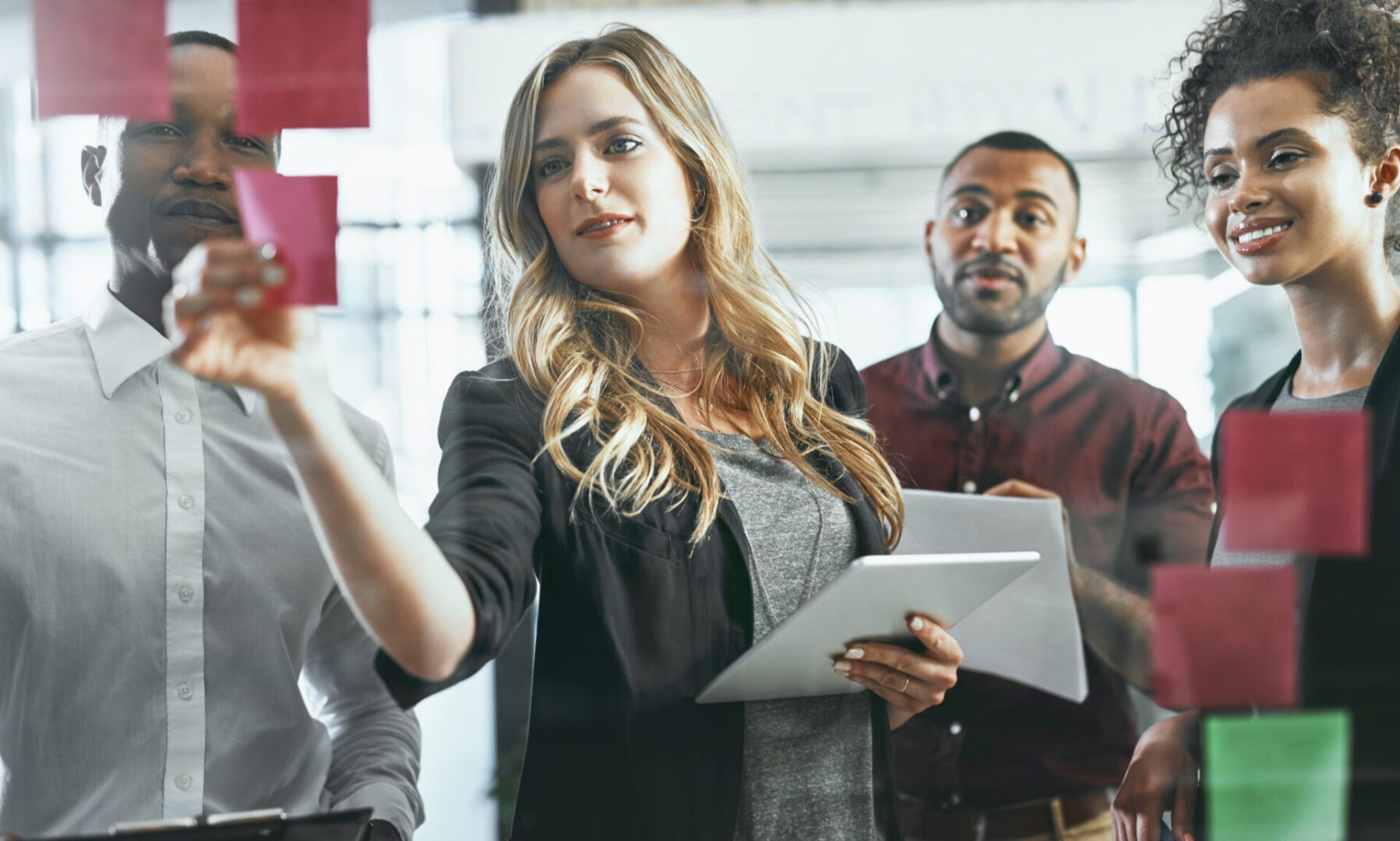 Group of professionals collaborating in an office, with a woman placing sticky notes on a glass wall while holding a tablet, surrounded by teammates.