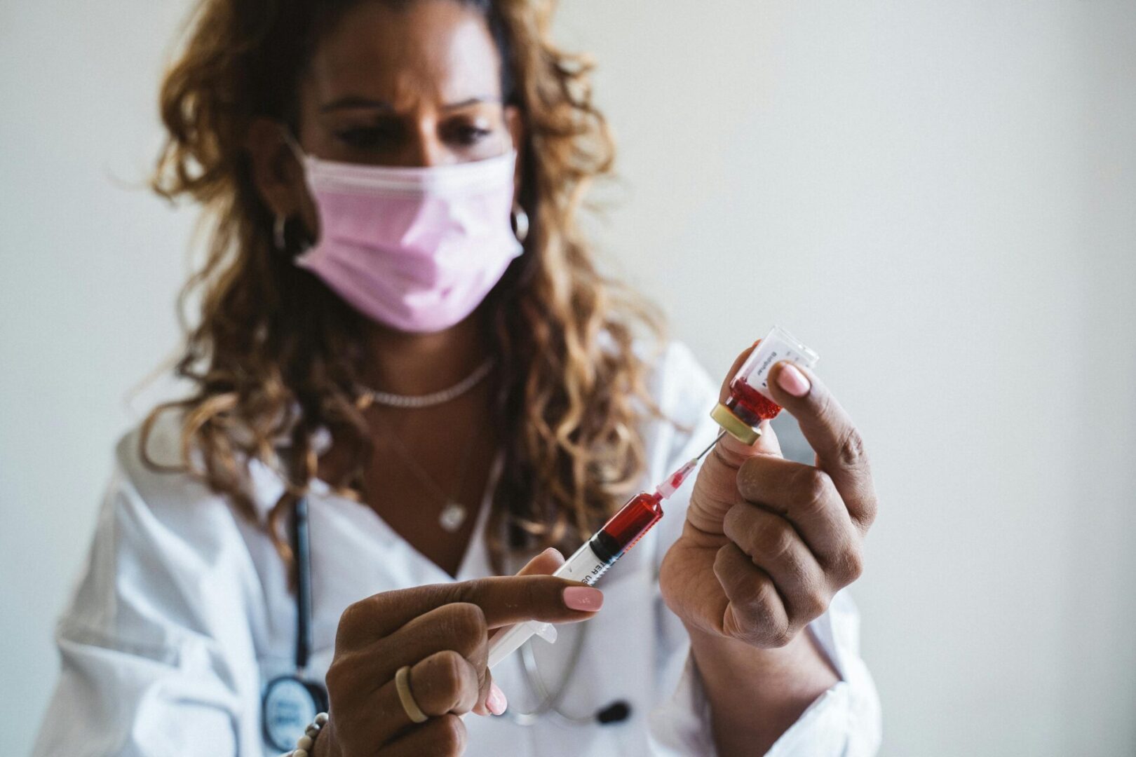 Female doctor wearing a pink face mask drawing red liquid into a syringe from a vial in a clinical setting.