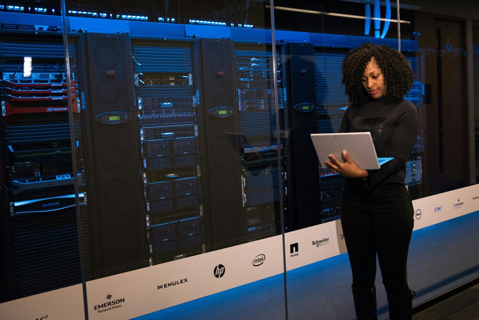 Woman standing in a server room, holding and working on a laptop in front of several server racks.