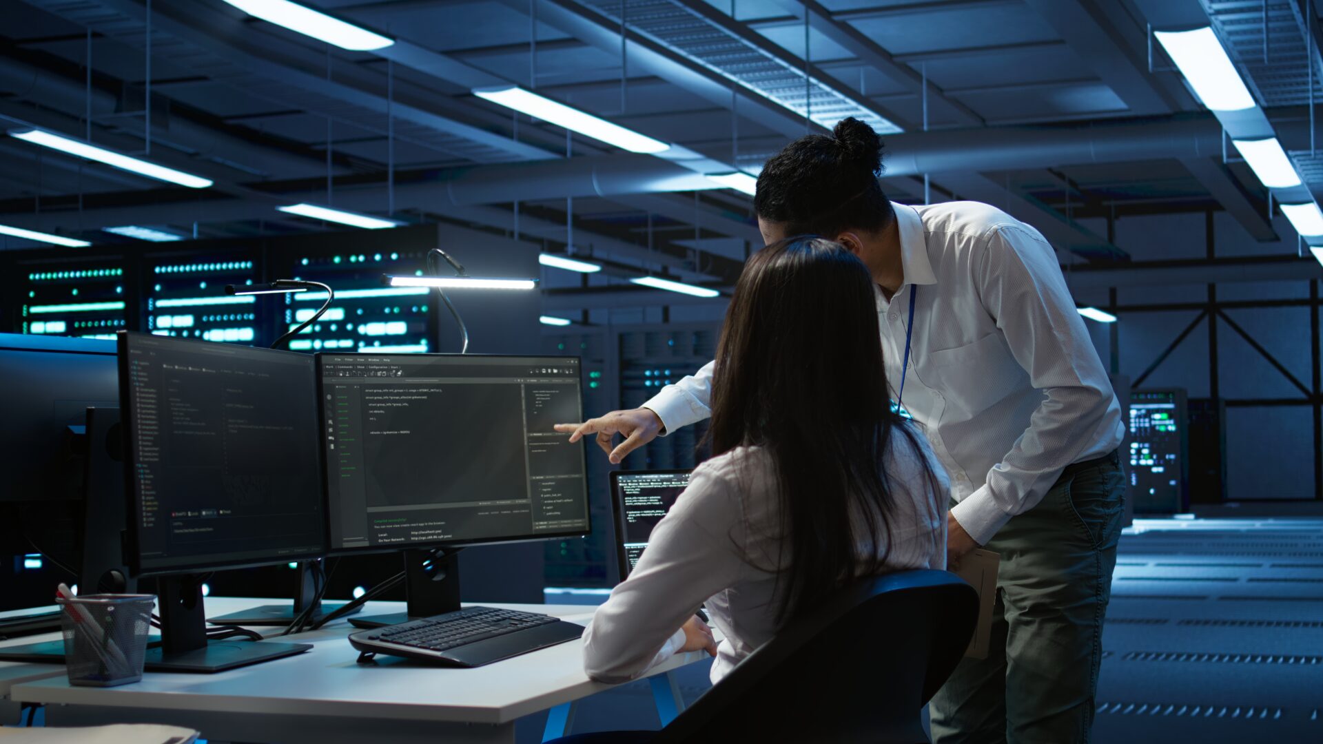 Two IT specialists in a dim, blue-lit data center: a woman sits at a desk with dual monitors showing code while a colleague leans over, pointing at the screen; rows of server racks with blinking lights fill the background.