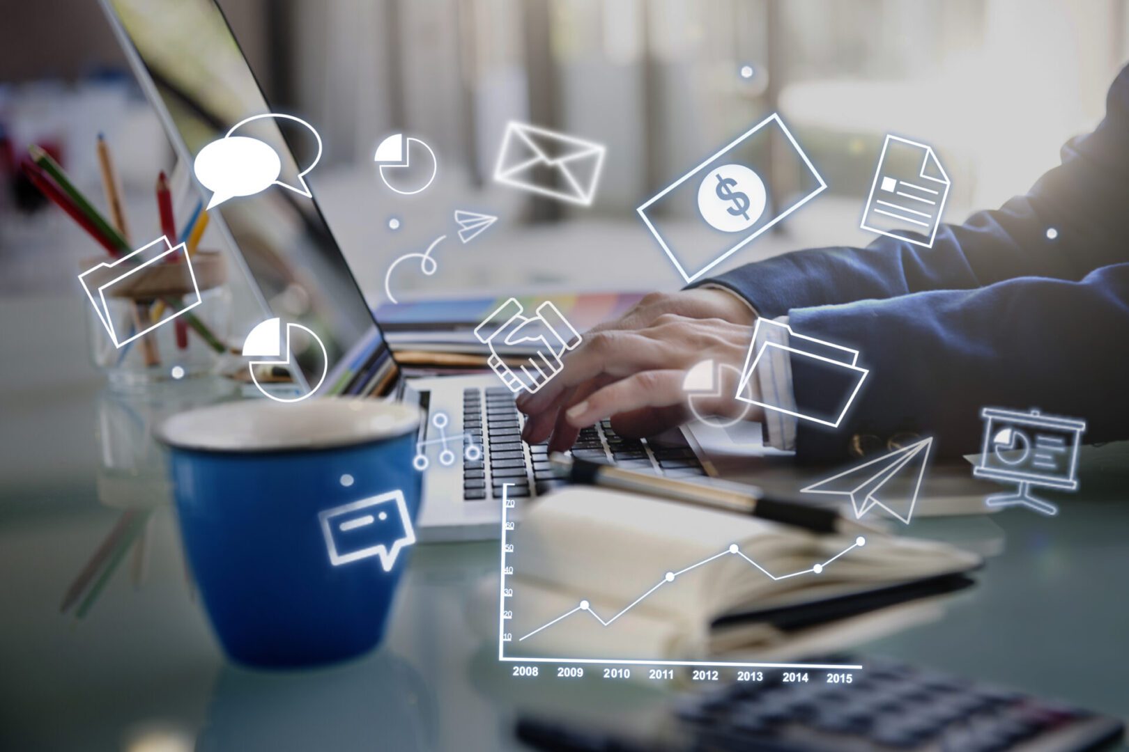 Hands of a person in a suit typing on a laptop at a tidy desk, with a blue coffee mug, notebook, and pencils nearby; translucent overlay icons of charts, dollar bill, envelope, folders, handshake, and line graph illustrate digital business and finance concepts.
