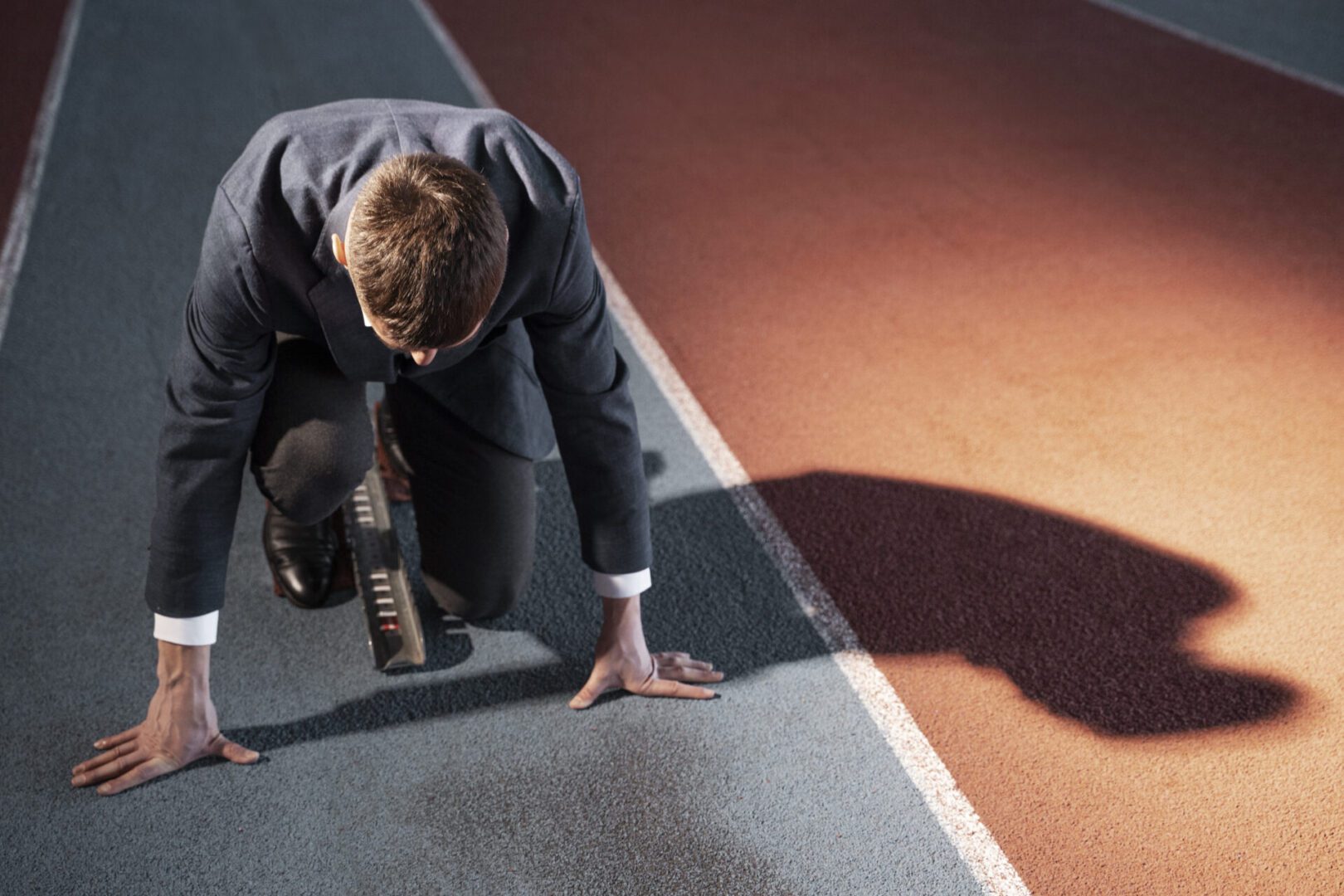 The image depicts a man dressed in a business suit crouched in a sprinter's starting position on a running track. He appears ready to launch into a race, blending the symbolism of athletic competition with professional ambition. The contrast between his formal attire and the athletic setting suggests themes like business competitiveness, drive, or the "race" for success in a corporate environment. The lighting casts a sharp shadow, adding a dramatic effect to the scene.