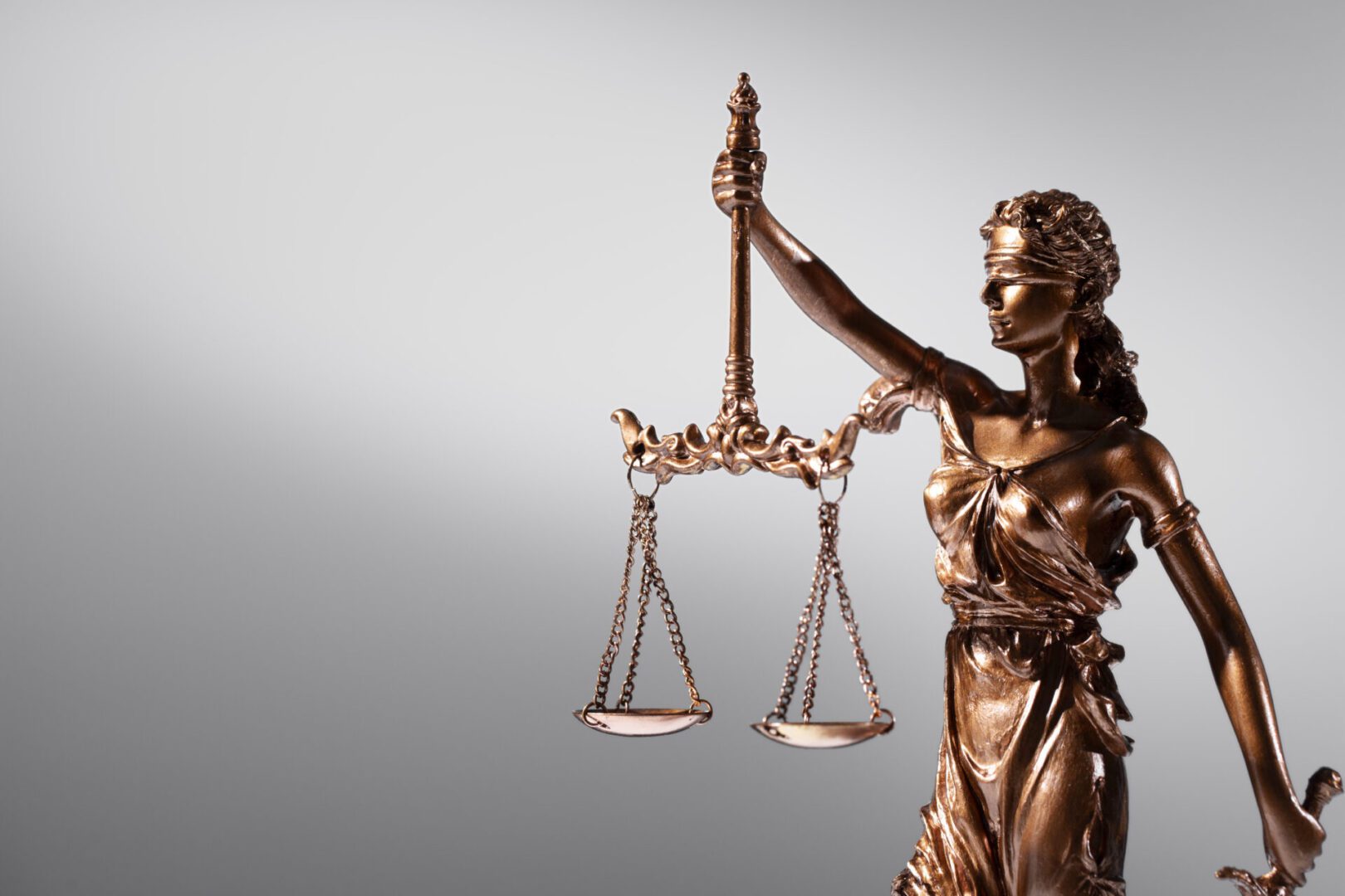 Bronze, blindfolded Lady Justice figurine on the right side of a soft gray gradient background, holding aloft a delicate set of balance scales while a sheathed sword rests at her side.