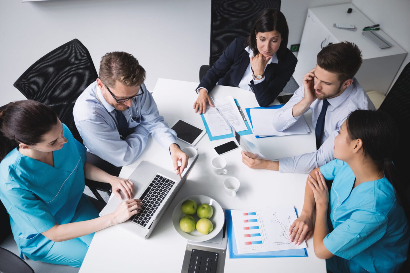 A group of healthcare and business professionals are having a meeting around a table, reviewing documents, charts, and a laptop. The setting looks collaborative and focused.
