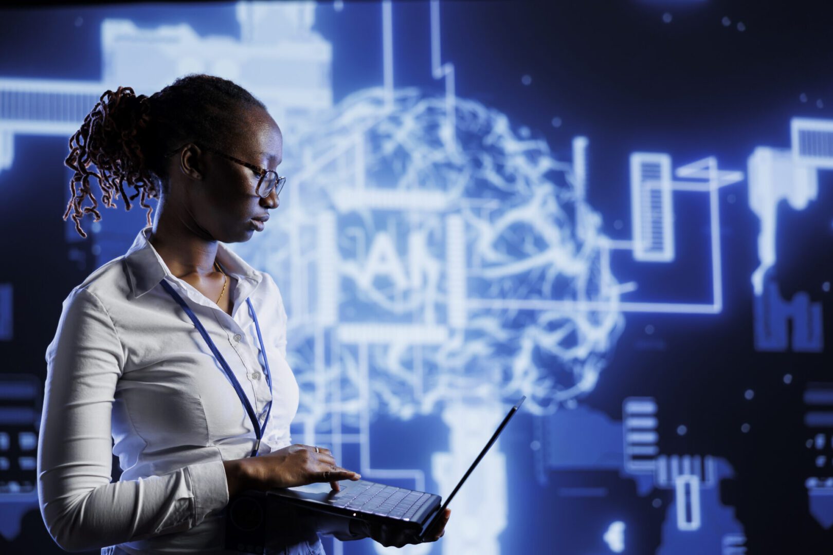 The image shows a woman wearing glasses and a white shirt, working on a laptop. Behind her is a large digital screen featuring a glowing blue brain graphic, representing artificial intelligence (AI) or a neural network, with circuit-like elements surrounding it. The overall environment and visuals suggest a high-tech or futuristic setting, possibly related to AI development, research, or data analysis.
