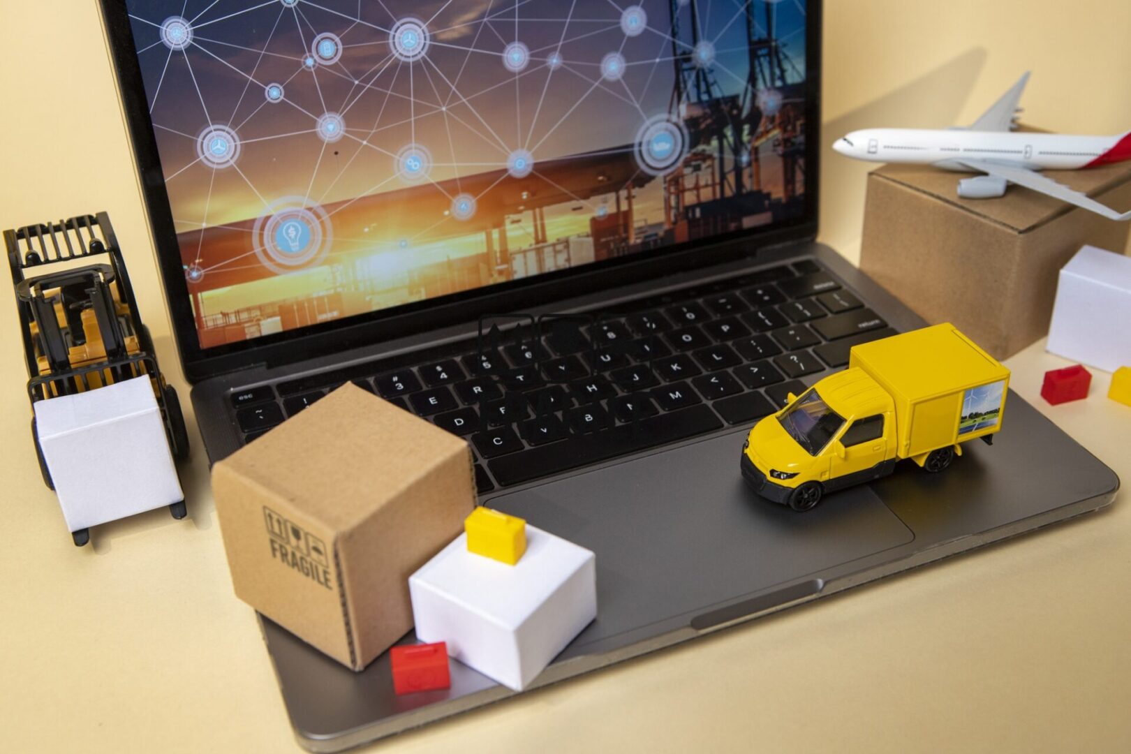 The image depicts a laptop with logistics-themed toys and props around it—such as a forklift, delivery truck, airplane, and boxes—symbolizing supply chain and logistics management through digital technology.