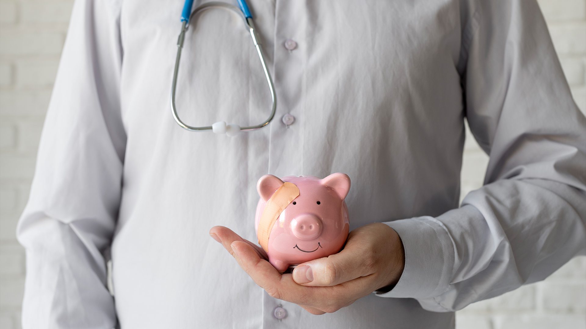 A person wearing a light gray shirt and a stethoscope around their neck is holding a pink piggy bank with a bandage on its forehead. The piggy bank has a small smile painted on its face, symbolizing financial health or medical expenses. The image conveys a concept related to healthcare costs, financial recovery, or saving money for medical needs.