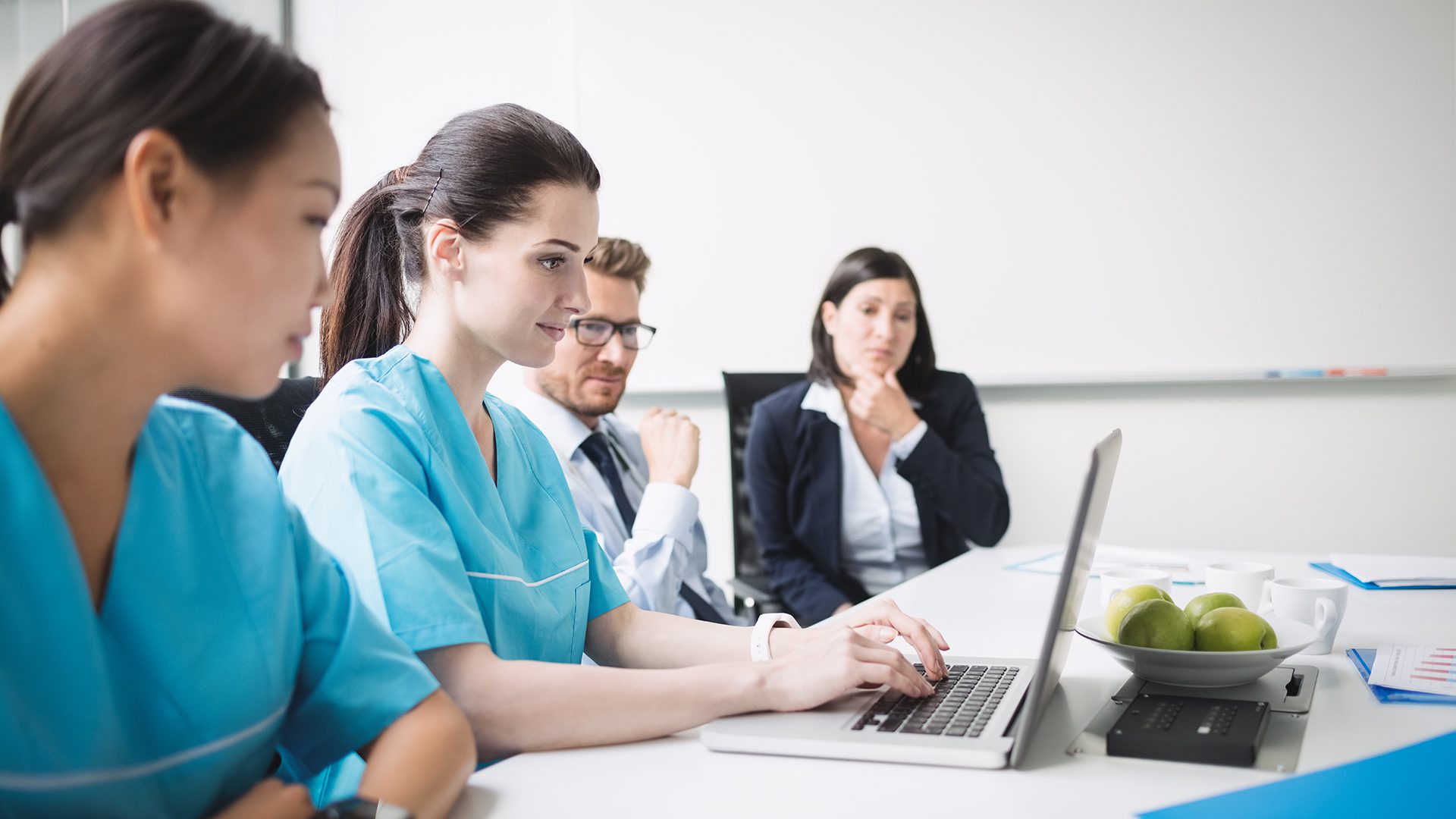 Four people, including two in medical scrubs, sit at a conference table. One types on a laptop. A bowl of apples is on the table. The group appears engaged in a meeting or discussion.