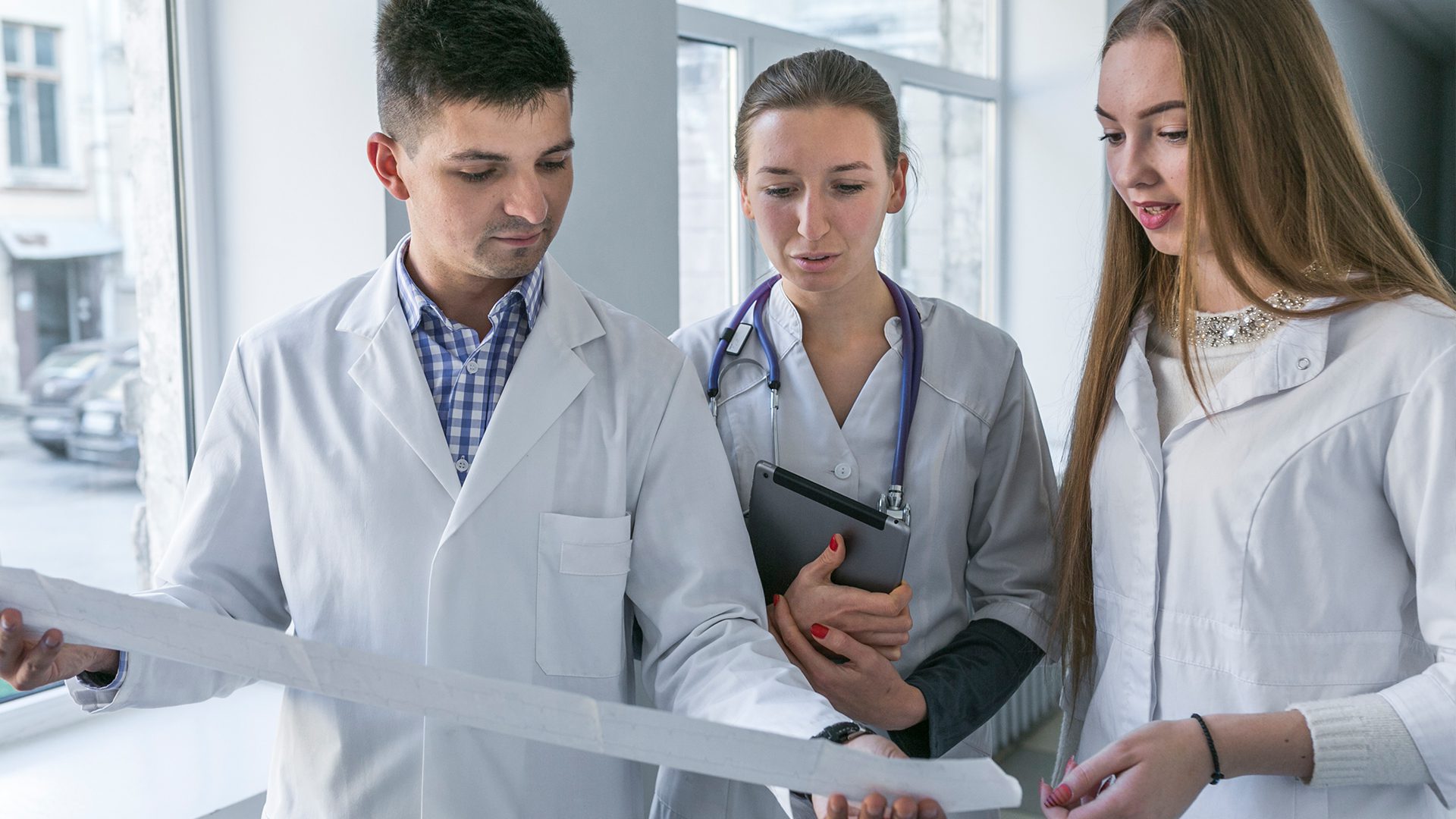 Three medical professionals, in lab coats, discuss a document while standing in a bright, modern hallway with large windows.