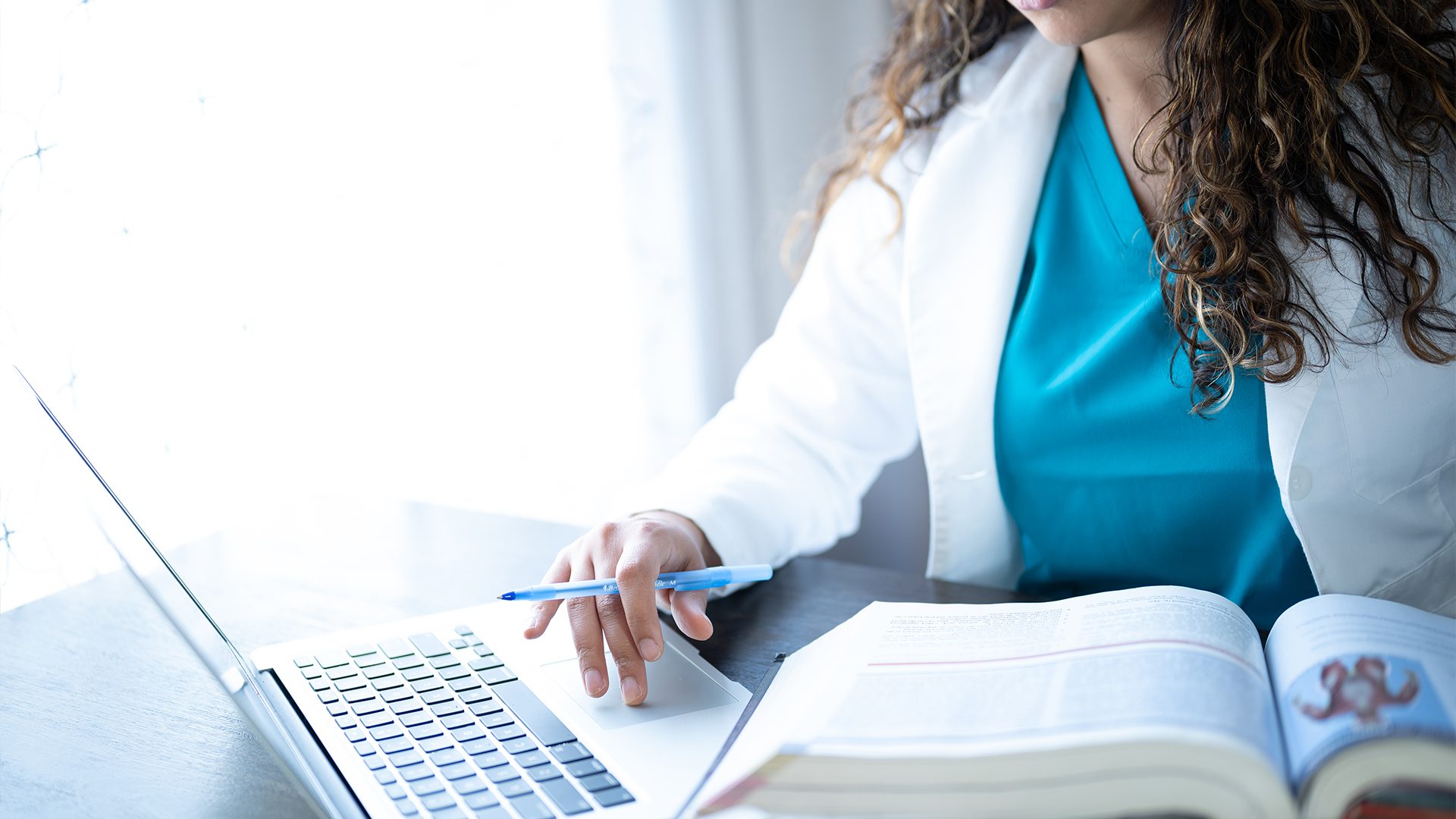 A healthcare professional in a white coat studies a laptop and medical book, holding a pen, focused on research or patient information.