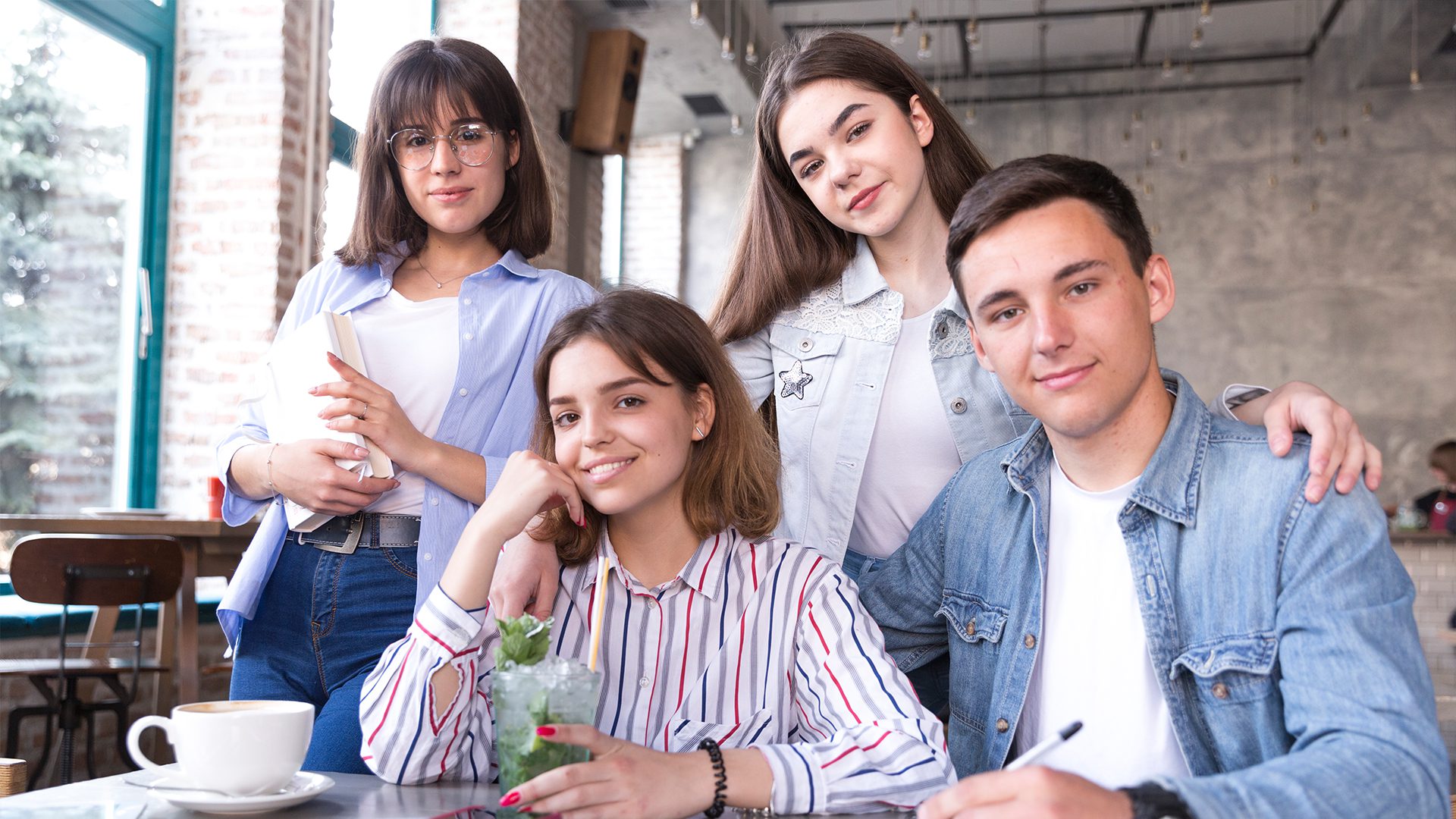 A group of four friends poses together in a cozy café, enjoying drinks and snacks, with bright windows and stylish decor in the background.