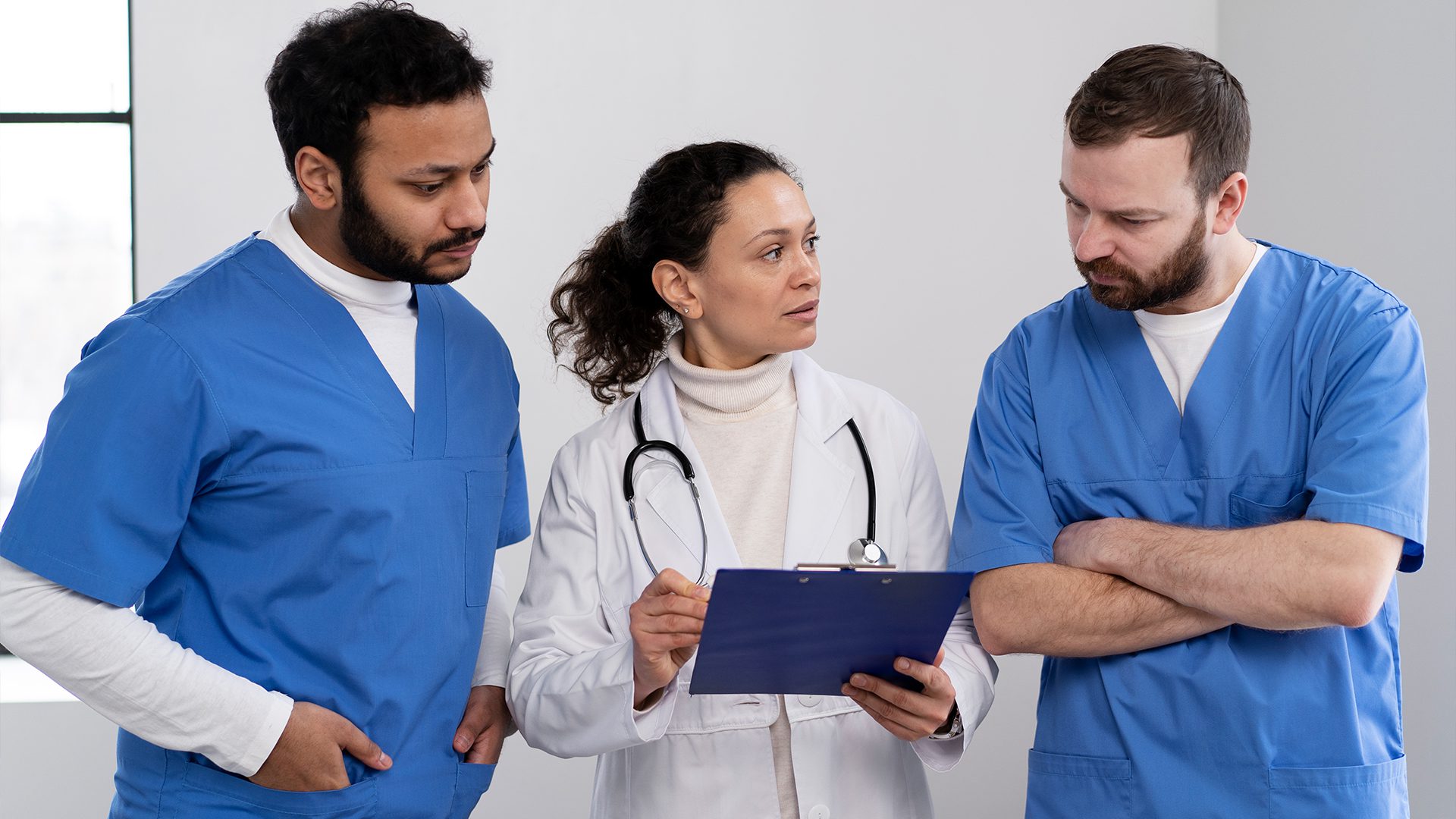 The image shows three medical professionals in discussion. The person in the center, wearing a white lab coat and a stethoscope, is likely a doctor or supervisor, holding a clipboard and speaking to the two individuals in blue scrubs on either side. The individuals in blue scrubs appear attentive and engaged, suggesting a collaborative or instructional moment in a clinical setting. The environment looks like a modern medical facility, possibly a hospital or clinic.