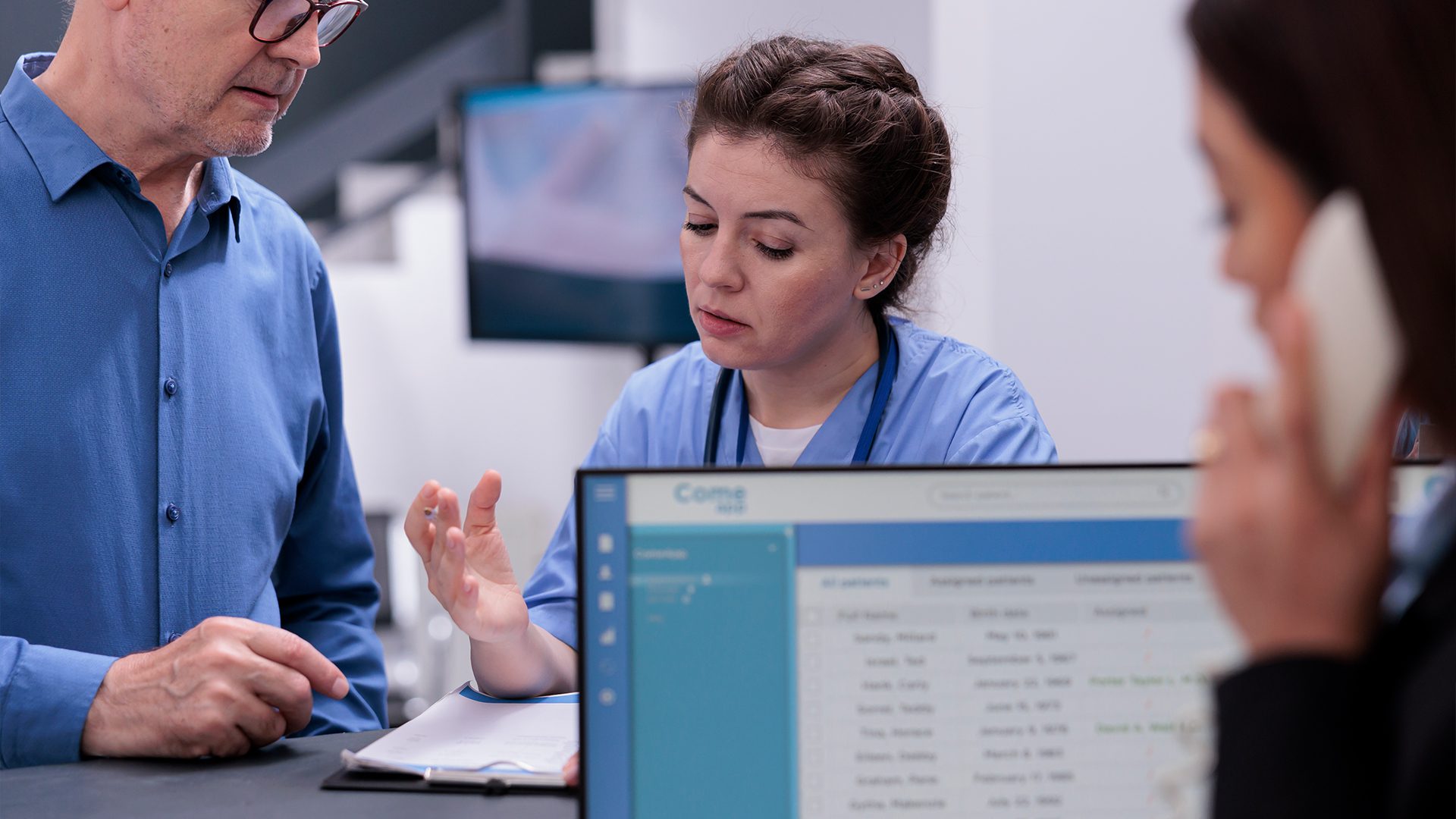 A nurse in blue scrubs is seated at a desk, attentively discussing information with an older man in a blue shirt. She gestures with her hand while looking at a clipboard. In the foreground, a woman is on the phone, and part of a computer screen displaying patient information is visible. The setting appears to be a hospital or clinic reception or nurse’s station. The atmosphere suggests a focused conversation, likely related to patient care or scheduling.