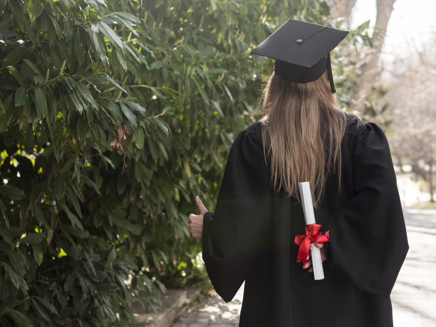 Rear view of a graduate in black cap and gown outdoors, holding a rolled diploma tied with a red ribbon behind her back and giving a thumbs-up gesture.