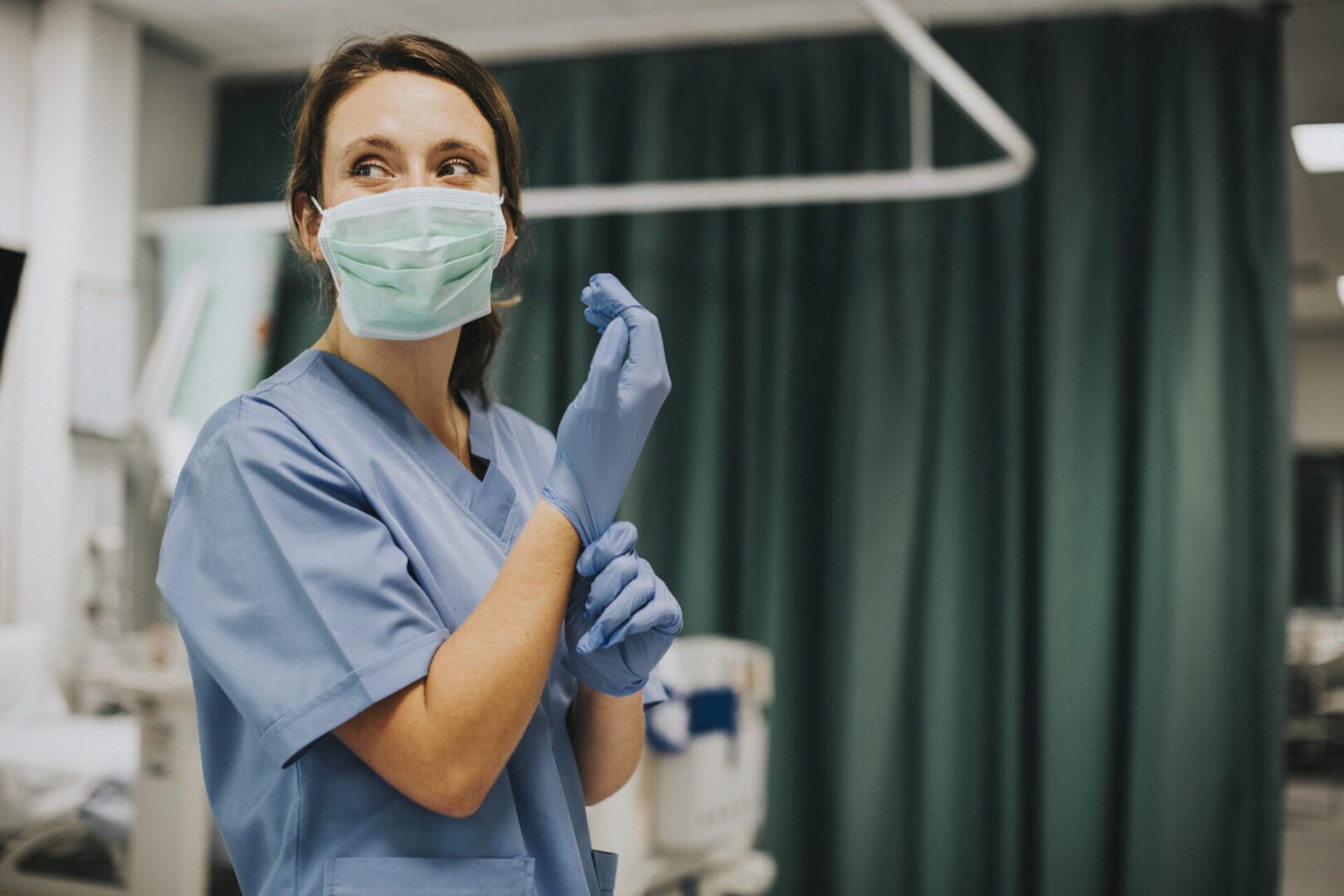 It looks like a healthcare professional—most likely a nurse—standing in a clinical setting. She’s wearing light-blue scrubs and a surgical mask, and she’s in the middle of putting on a pair of exam gloves. Behind her you can see a green privacy curtain and some blurred medical equipment, giving the impression this is an exam room or ward.