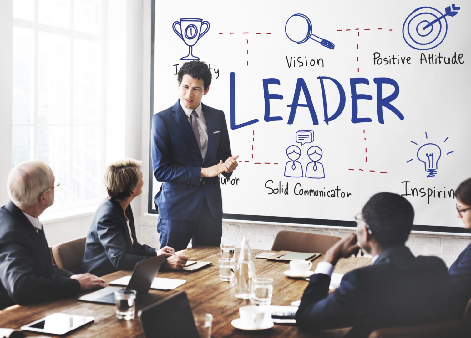 Business meeting where a young man in a blue suit stands by a whiteboard covered with the word ‘LEADER’ and hand-drawn icons like a trophy, magnifying glass, target, and lightbulb, explaining leadership traits to seated colleagues around a conference table.