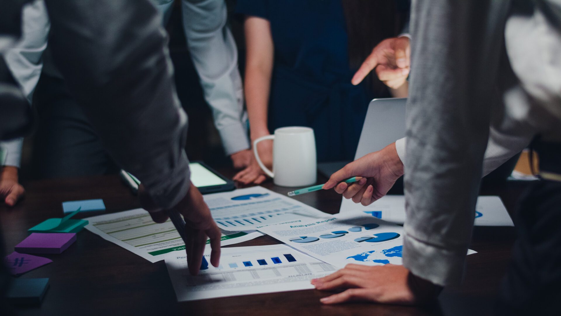 Group of young Asian professionals gathered around a dimly lit office table, pointing at printed charts, bar graphs, and pie charts during a collaborative nighttime brainstorming session.
