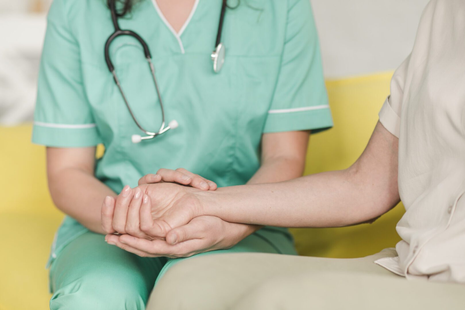 Nurse in teal scrubs, stethoscope around neck, gently holding and checking the pulse at a seated female patient’s wrist.