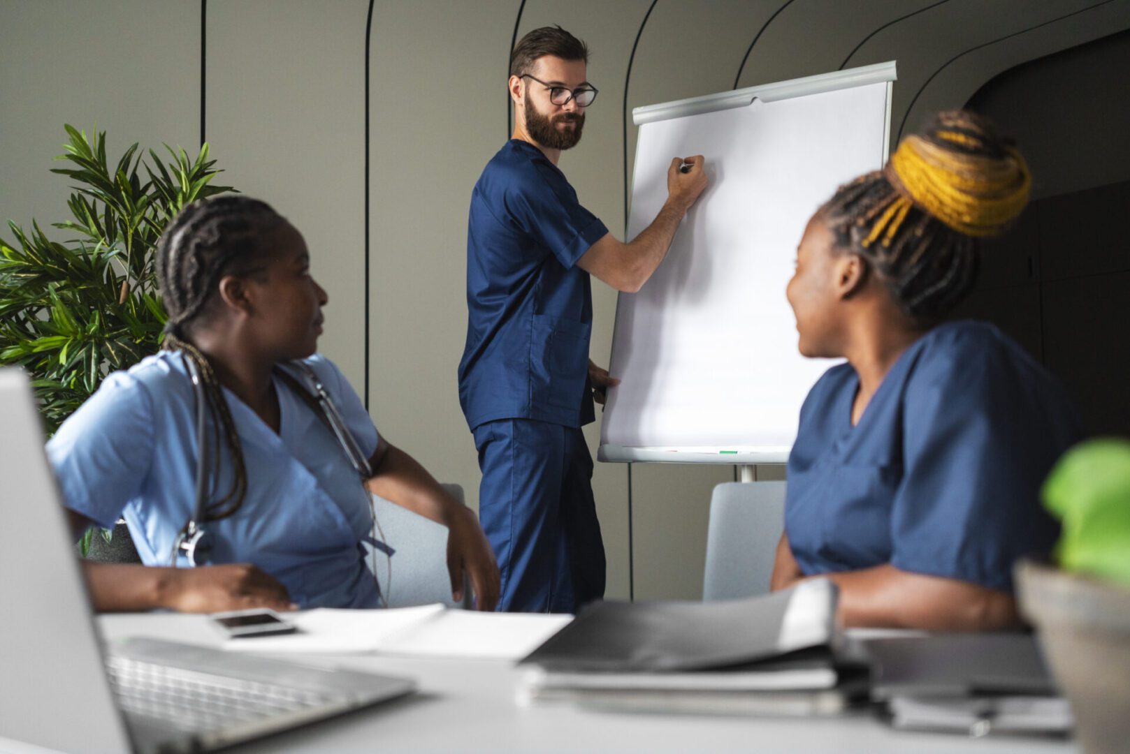 Three nurses in blue scrubs, one standing and writing on a flip-chart while two others sit at a table with notebooks and a laptop during a training session.