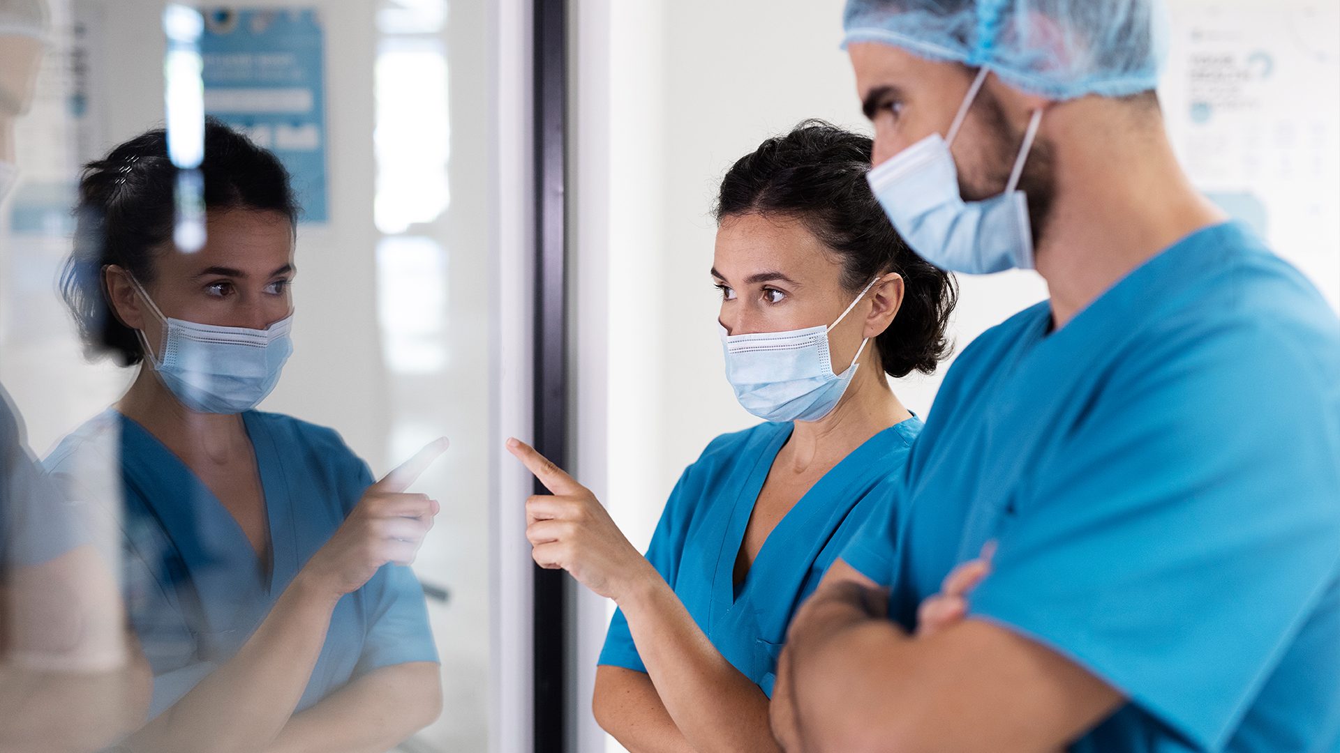 Two healthcare professionals in blue scrubs and masks stand indoors, looking at and pointing toward something outside the frame through a glass window.