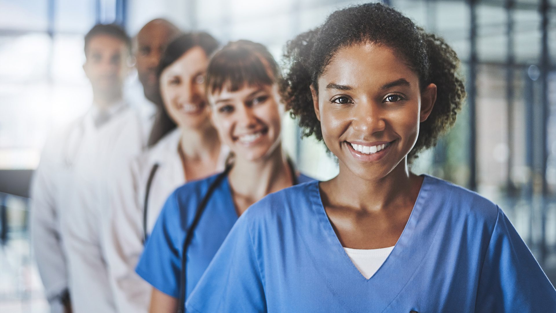 A group of healthcare professionals, including men and women in scrubs and lab coats, stand in a line and smile at the camera in a clinical setting.