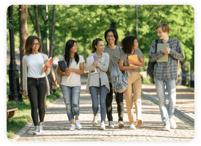 A diverse group of six students walks along a tree-lined pathway in a park-like setting, chatting and smiling. They are casually dressed and carrying books, notebooks, and backpacks, suggesting a school or college environment. Sunlight filters through the green trees, creating a warm and inviting atmosphere